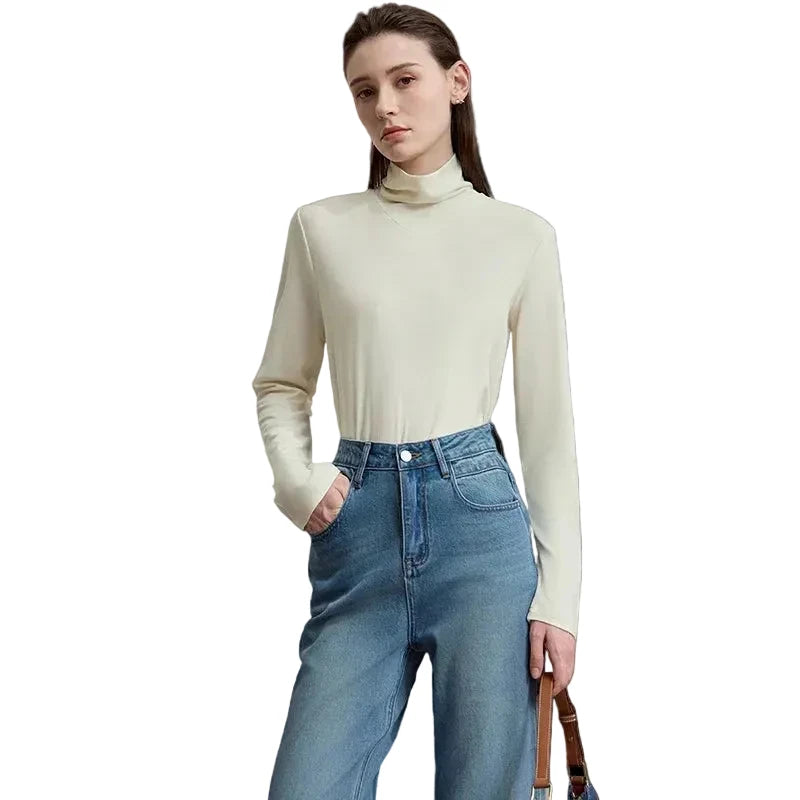 A woman modeling a fitted, long-sleeve cream mock-neck top paired with high-waisted blue jeans, standing against a neutral light gray studio background. The brand name 'AMII' is printed in black in the top-left corner.